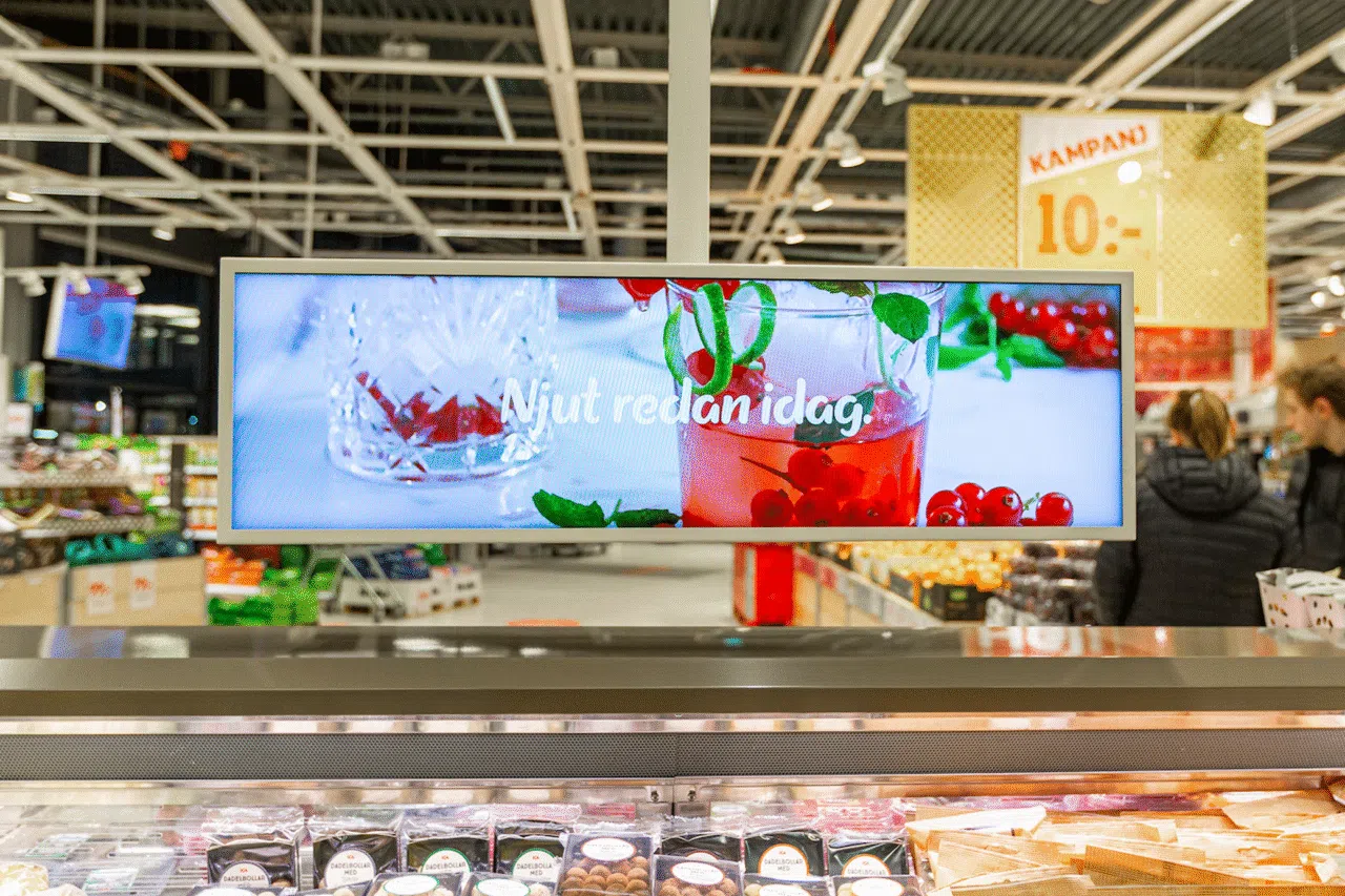 Digital display in a grocery store showing a glass of red berry drink with mint leaves and the Swedish text 'Njut redan idag' (‘Enjoy today’), placed above a refrigerated food section.