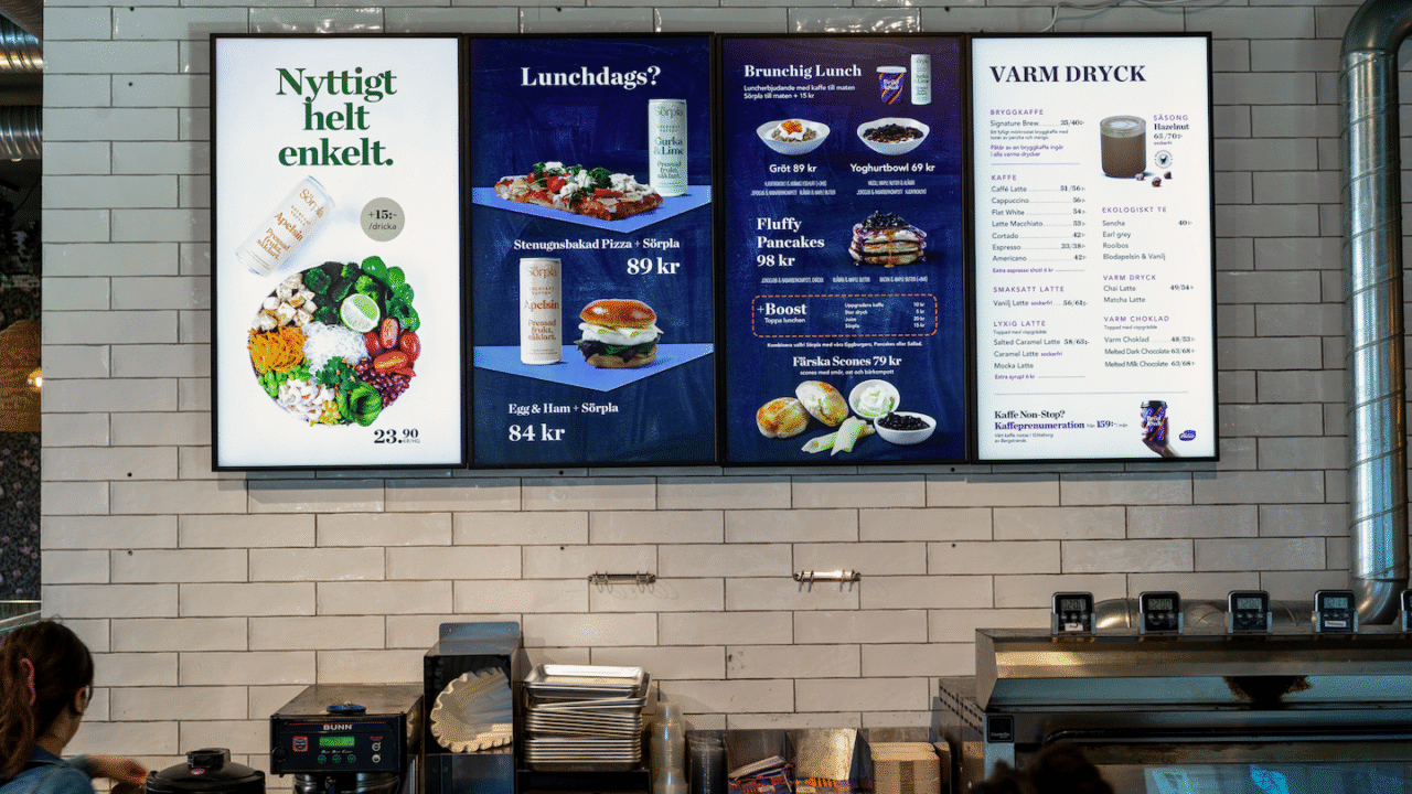 Digital menu board in a restaurant, showing colorful images of salads, smoothies, and lunch items with prices in Swedish, above a kitchen area.