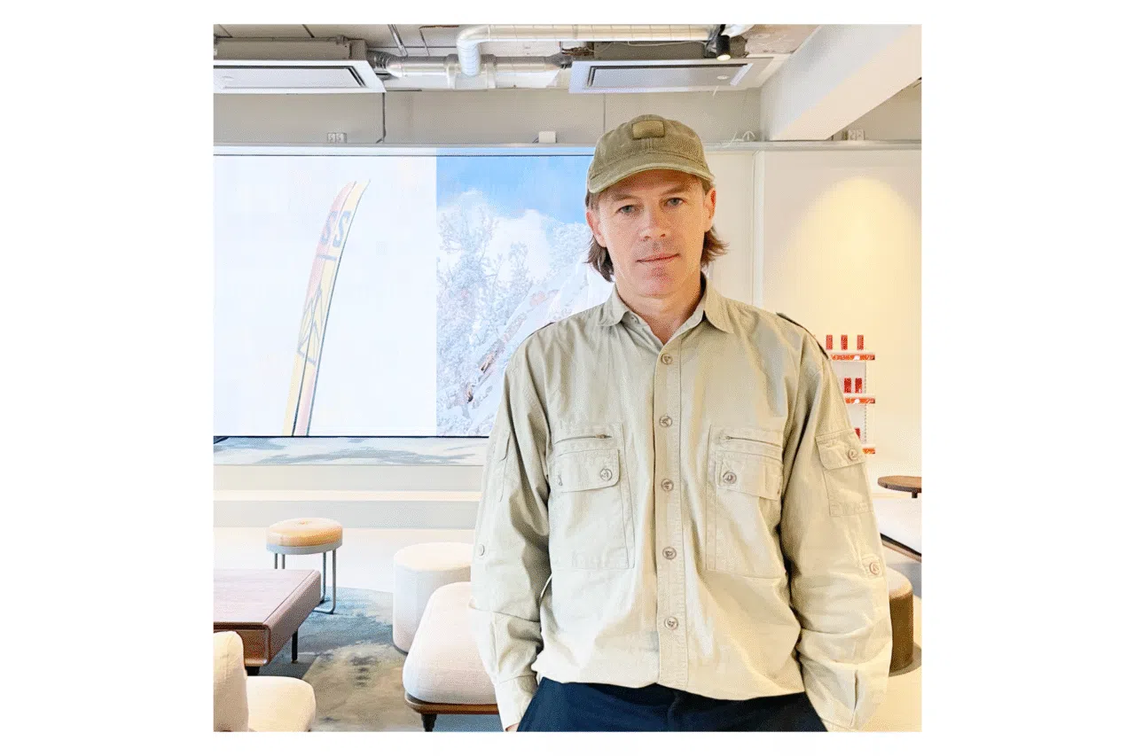 A person in a beige shirt and cap stands in a modern office space with cushioned stools. Background features a colorful abstract design on a LED screen. Professional, creative atmosphere.